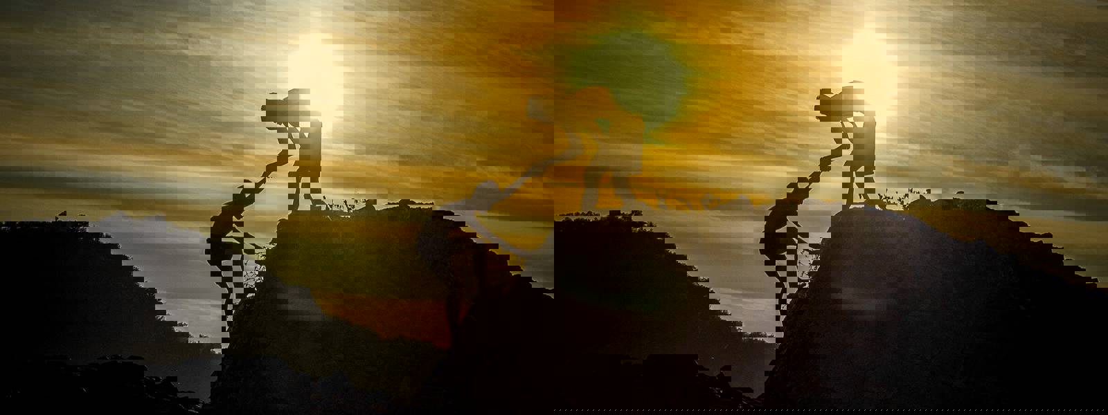 Two silhouetted figures helping each other climb a rocky outcrop against a sunset backdrop.