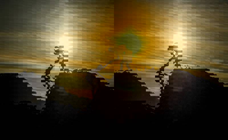 Two silhouetted figures helping each other climb a rocky outcrop against a sunset backdrop.