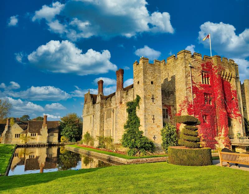 Historic Hever castle surrounded by greenery and a reflective pond under a blue sky.