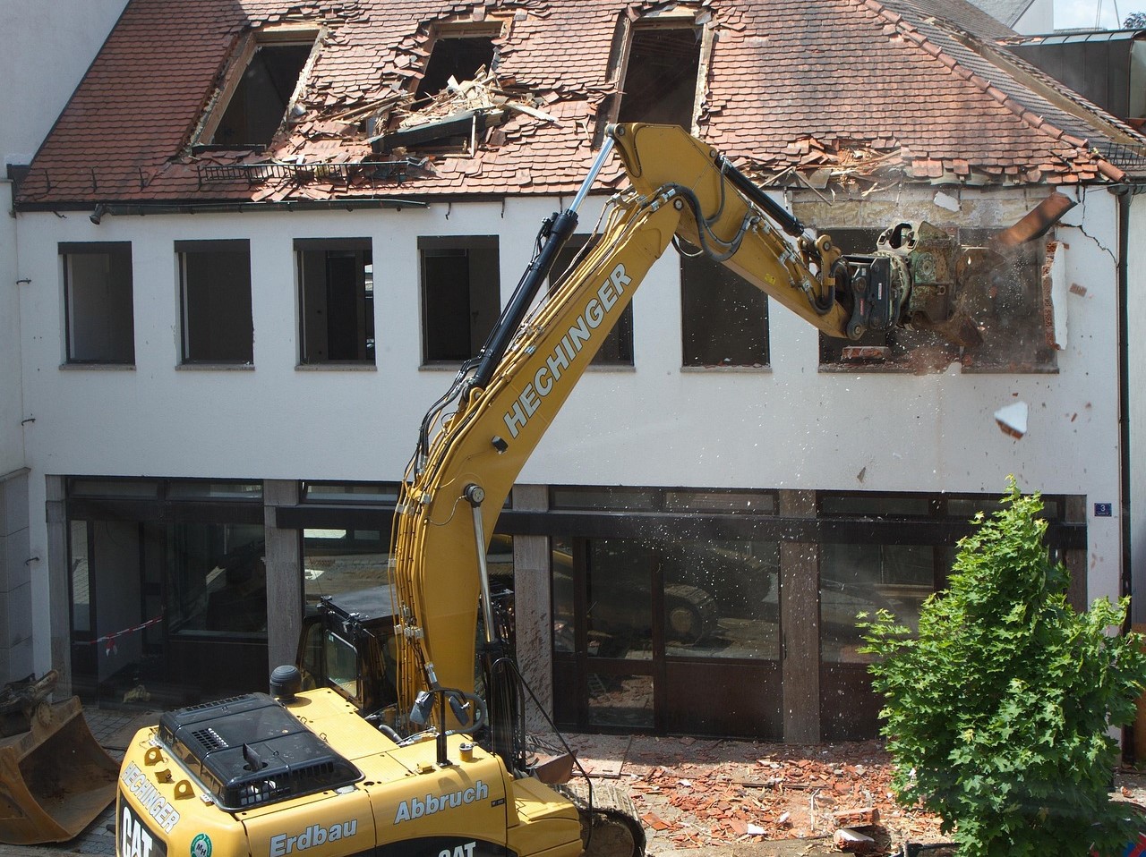 Excavator demolishing a building, debris falling from the roof in a construction site.
